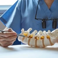 Doctor in blue scrubs demonstrating a spine model with a surgical instrument.