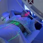 Patient lying on a treatment table in a medical imaging or radiation therapy machine, with laser alignment lines projected onto their body.