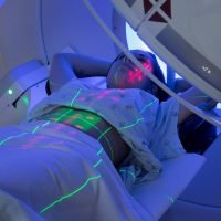 Patient lying on a treatment table in a medical imaging or radiation therapy machine, with laser alignment lines projected onto their body.